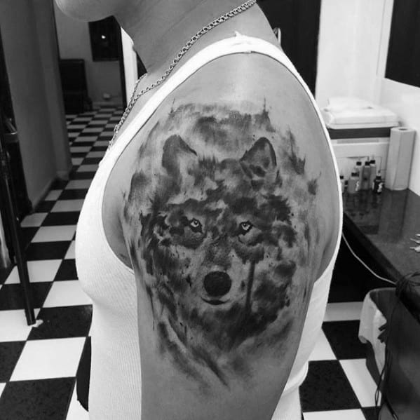 Black and grey wolf tattoo on upper arm with smoky shading and piercing eyes, blending realism with a soft, mist-like texture.