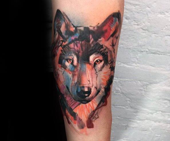 Watercolor wolf tattoo with vivid red, blue, and black tones, soft brushstrokes, and a calm yet powerful gaze on the forearm.