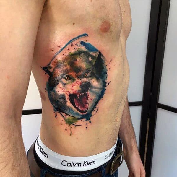 Ferocious wolf tattoo on the side of the torso with mouth open mid-snarl, enhanced by bold watercolor splashes in blue, red, and green.