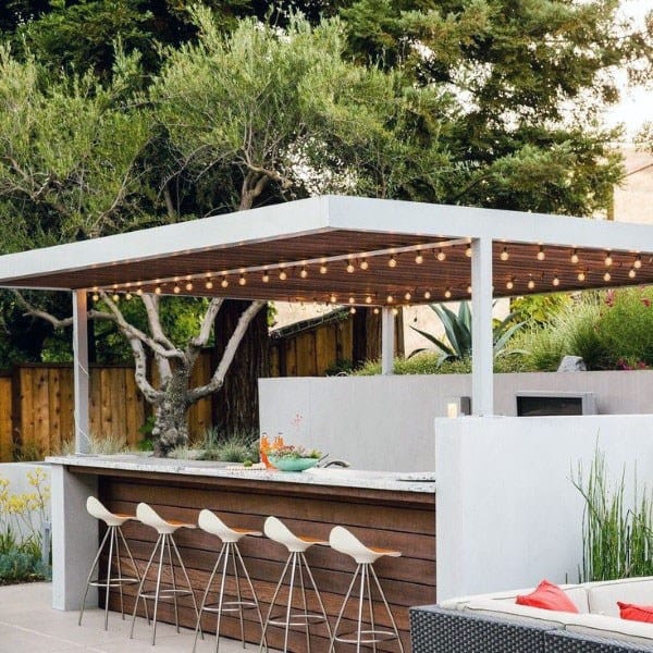 Minimalist backyard bar with string lights, white stools, and wooden accents under a modern pergola.