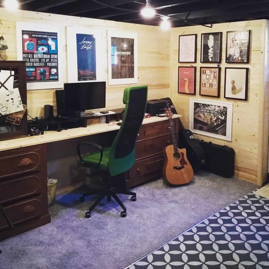 Music room/office with wooden walls, a desk, green chair, guitar, and framed posters on the wall.
