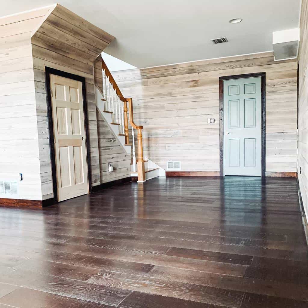 Spacious living room with light wood-paneled walls, dark hardwood floors, and a wooden staircase.