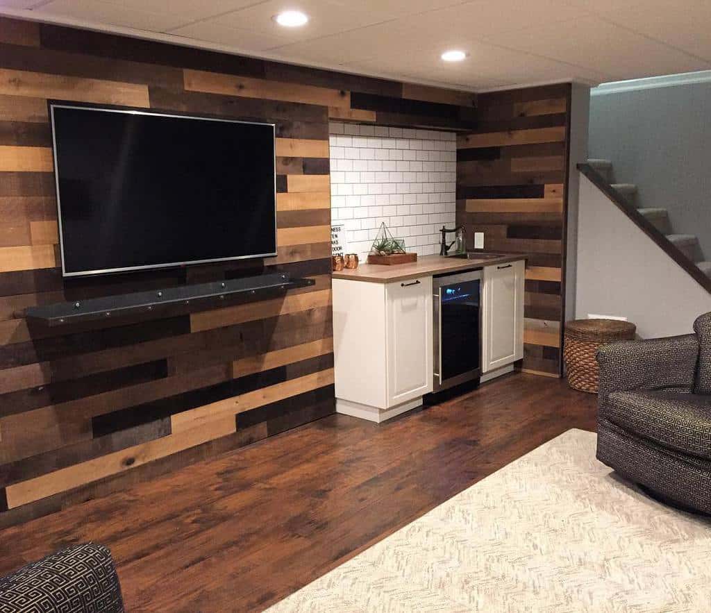 Basement living room with wood accent wall, wall-mounted TV, and small white kitchenette.