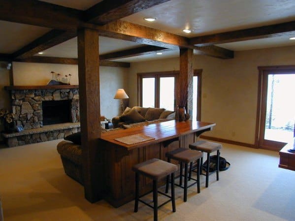 Rustic basement with wooden beams, a stone fireplace, bar seating, and cozy lounge furniture.