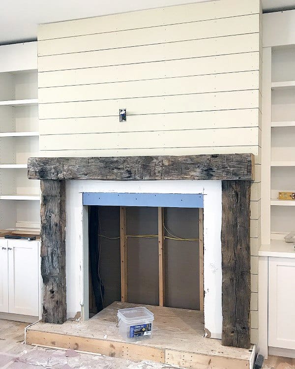 Rustic fireplace with reclaimed wood beams, shiplap wall, and built-in shelves under construction.