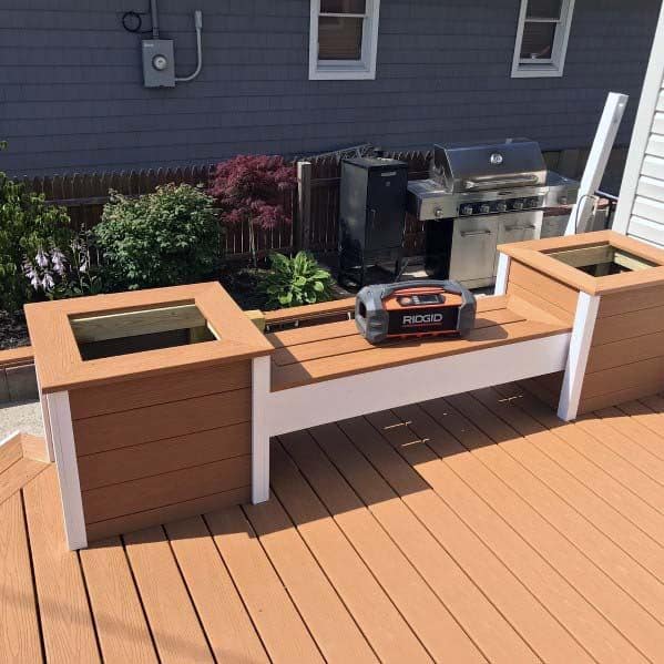 Wooden deck with planter boxes, a portable radio, and a bench by the stainless steel grill near windowed wall