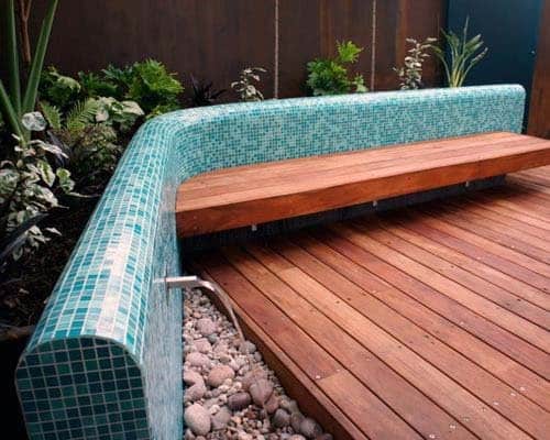 A wooden bench features a blue tiled backrest, nestled on a wooden deck surrounded by lush plants and stones