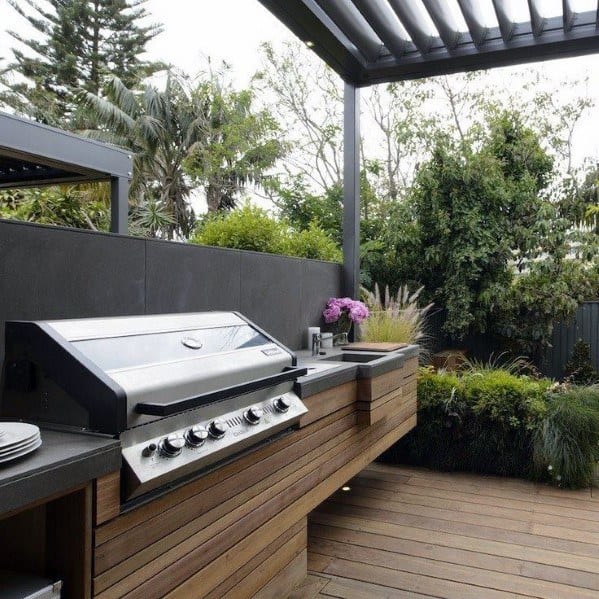 Outdoor kitchen with a built-in grill, wooden cabinets, and a lush garden backdrop