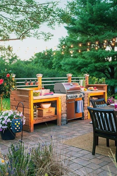 Outdoor kitchen with a built-in grill, brick patio, string lights, and surrounding greenery, cozy seating area in front