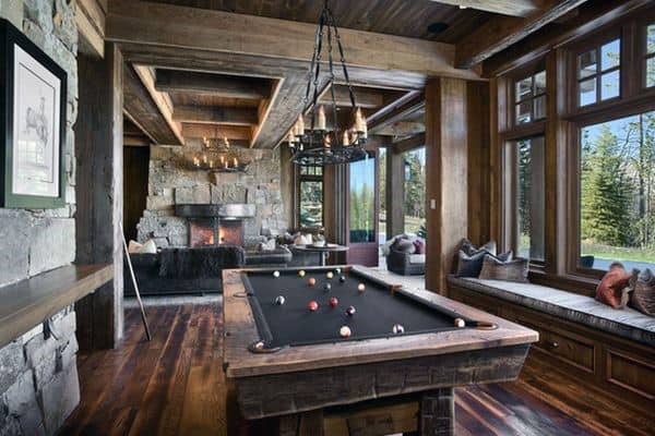 Rustic game room with a wooden pool table, stone fireplace, and large windows offering a view of trees