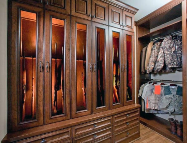 Custom wood cabinetry for gun storage with glass display doors, integrated lighting, and adjacent hunting gear closet.
