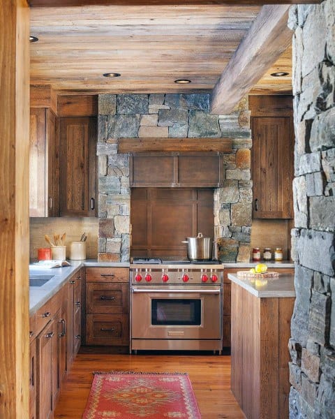 Rustic kitchen with stone walls, wooden cabinets, stainless steel stove, and a red accent rug.