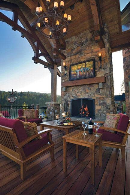 Cozy outdoor patio with wooden furniture, a stone fireplace, and a view of trees under a vaulted ceiling