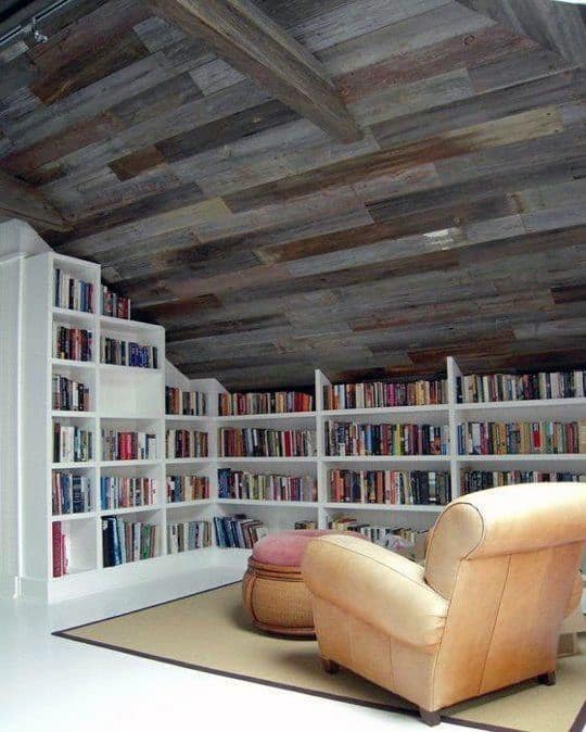 Cozy attic library with white bookshelves, a tan armchair, round stool, and wooden ceiling