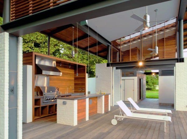 Modern outdoor kitchen with stainless steel appliances, wooden cabinetry, and two white outdoor lounge chairs