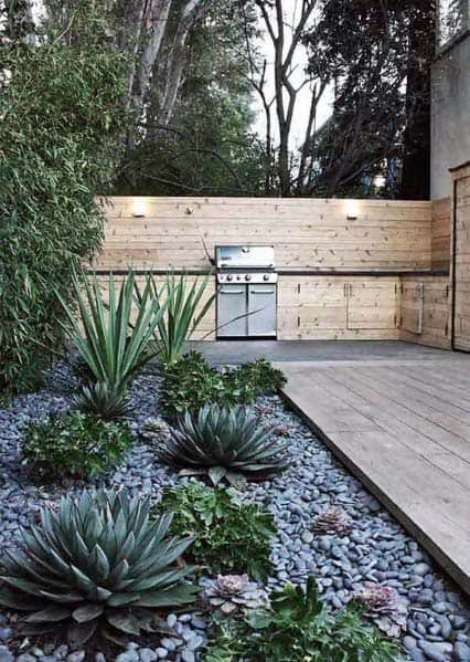 Stylish outdoor kitchen with a wood deck and river rock landscaping, featuring agave plants and a sleek grilling station