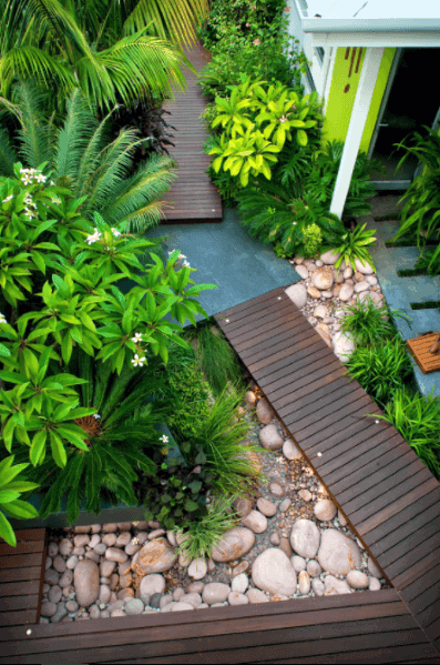 Wooden deck walkway with river rock landscaping, surrounded by lush tropical plants, creating a serene and stylish outdoor path