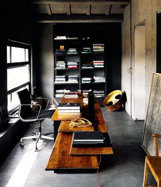 Minimalist office with wooden desk, black chair, large bookshelf filled with books, and a metal drum in the corner