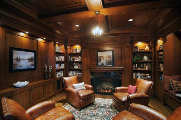 Cozy library with leather chairs, bookshelves, a fireplace, and framed art on wood-paneled walls