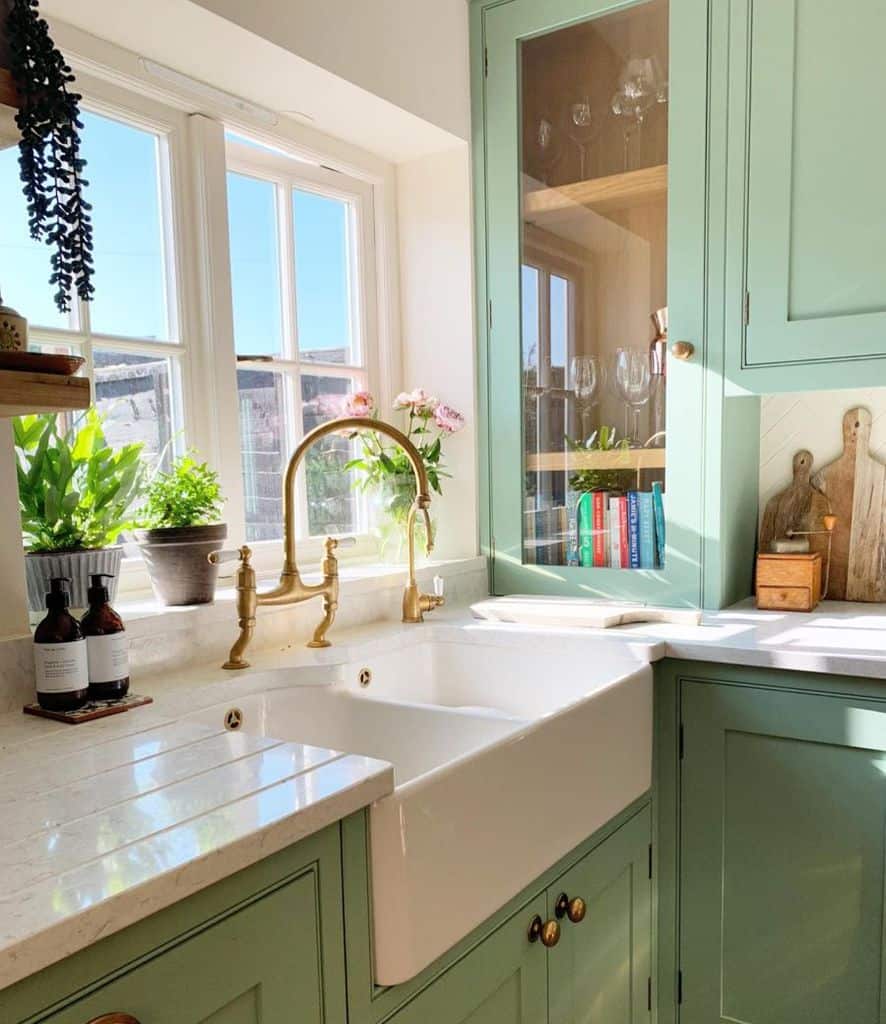 Charming kitchen with a farmhouse sink, brass faucet, green cabinetry, and a sunlit window.