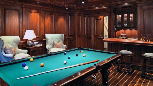 Cozy billiard room with wood paneling, armchairs, a green felt table, and a small bar area.