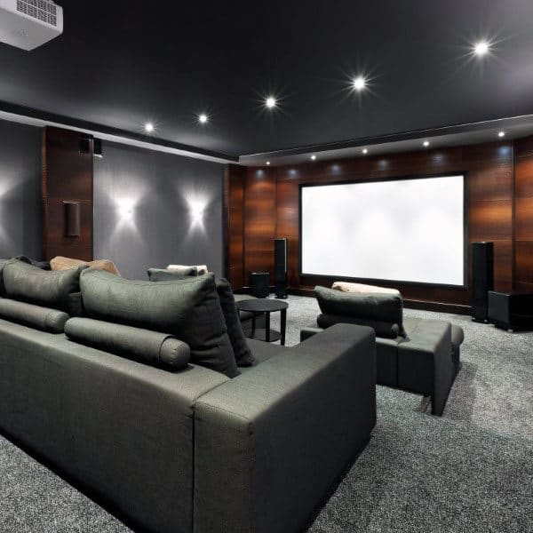 Home theater room with large screen, dark sofas, and surround sound speakers under dim ceiling lighting