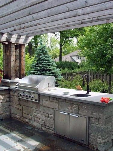 Outdoor kitchen with a built-in grill, sink, and stone countertops under a wooden pergola, surrounded by greenery
