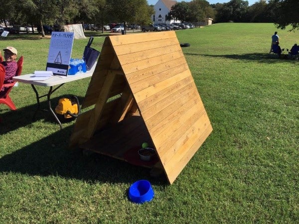 Simple wooden A-frame dog house with an open front, placed on grass with water bowls outside, set in a spacious outdoor area