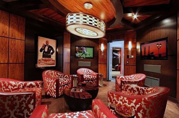 Cozy man cave with red patterned chairs, wooden walls, TVs, a chandelier, and posters decorating the room