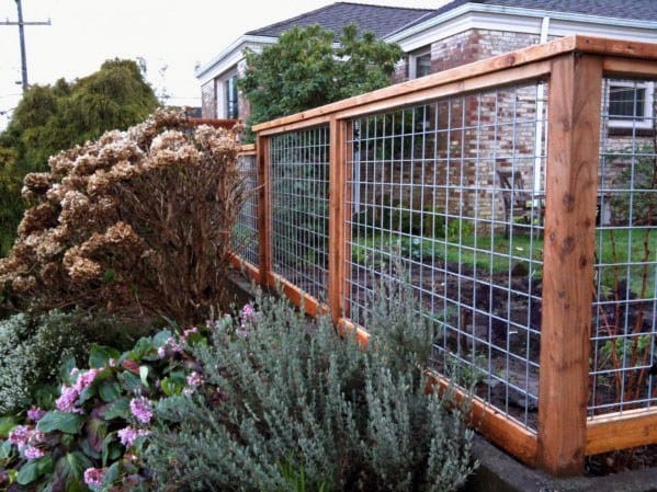 Garden with wooden fence, wire mesh, brick house backdrop, and an array of shrubs and flowers in the foreground