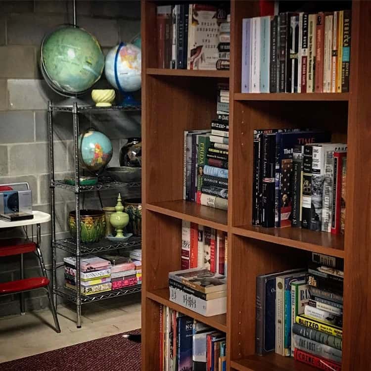 Wooden bookshelf in a basement, filled with various books and organized neatly next to a metal shelf holding globes and decorative items.
