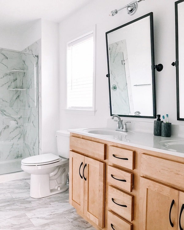 A modern farmhouse bathroom with a marble shower, wooden vanity, dual mirrors, and a white toilet under a bright window