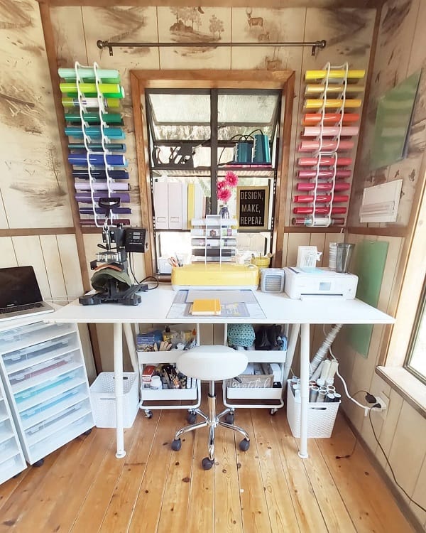 Bright craft room with a white desk, colorful paper racks, cutting machines, and supplies on wooden floors