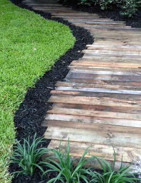 Walkway of wooden garden path with dark mulch, bordered by grass and small plants