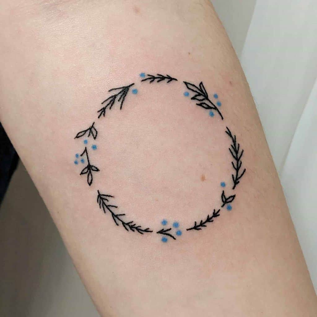 A simple and delicate tattoo of a circular wreath made of leaves and small blue flowers, placed on the forearm.