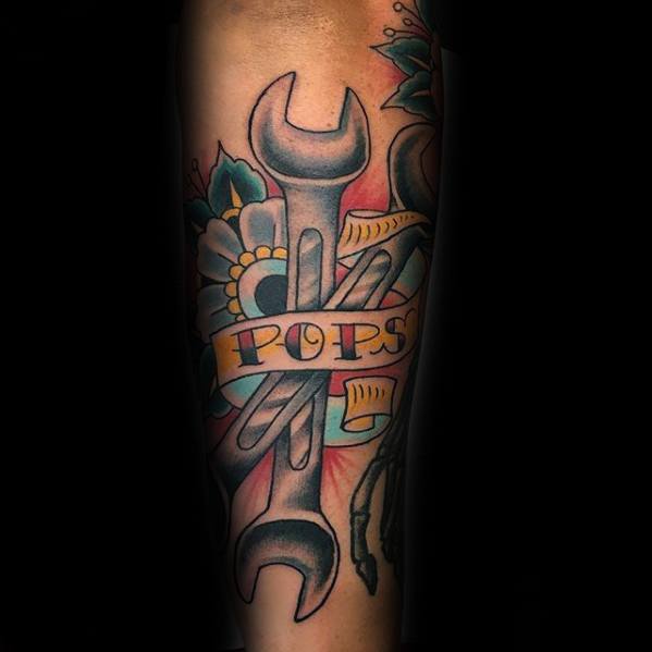 Colorful tattoo of two crossed wrenches with a yellow banner reading POPS, surrounded by flowers and barber tools in a traditional style.