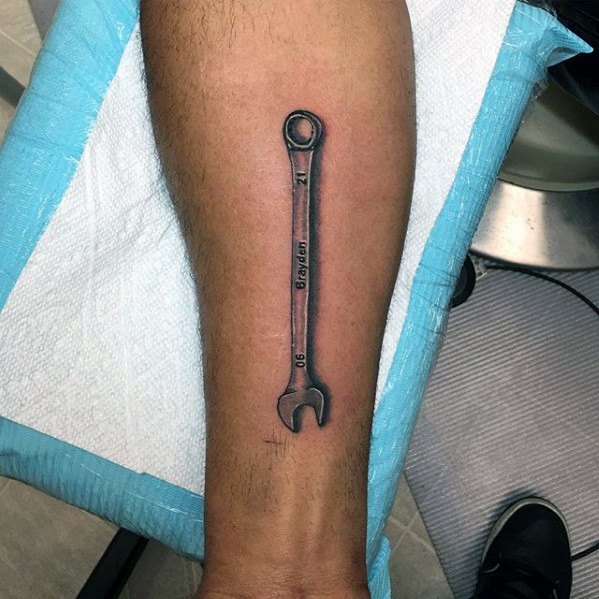Realistic tattoo of a long combination wrench on the leg, engraved with “Boylan,” “21,” and “06,” featuring soft shading for a 3D look.