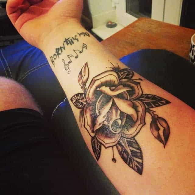 Forearm tattoo of a rose and leaves, with musical notes and "Born this way" text above