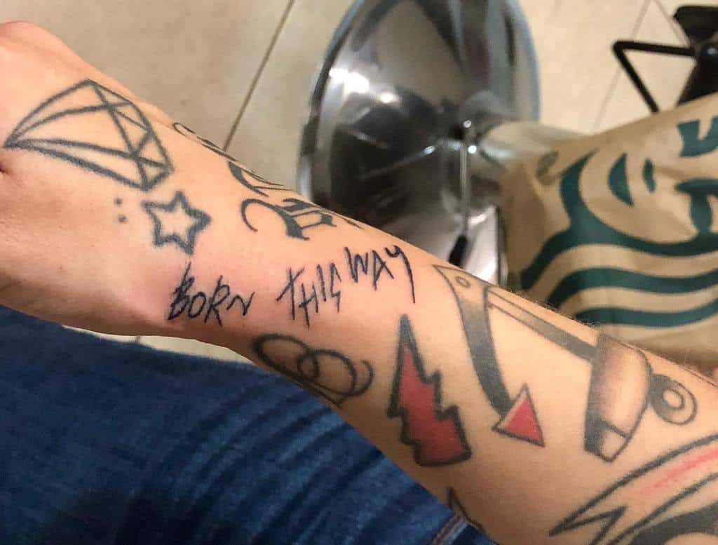 Arm with various tattoos including a diamond, star, safety pins, and "Born This Way" text