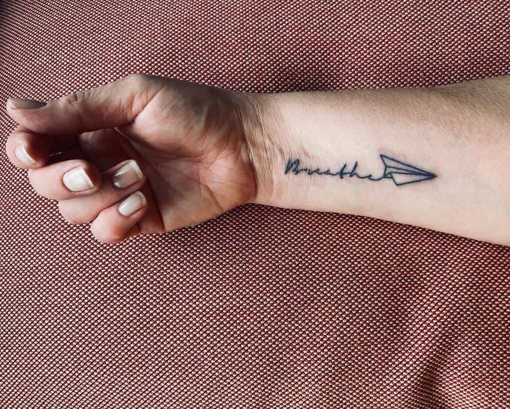 A wrist tattoo elegantly features a paper airplane soaring above the word "breathe," etched delicately on the wrist