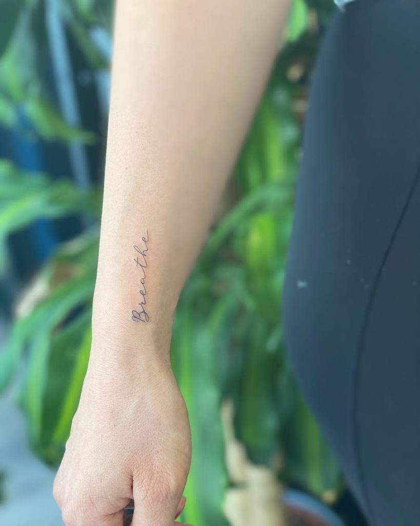 A close-up reveals a forearm graced with an elegant "Breathe" tattoo in cursive