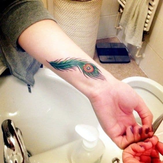 Small peacock feather tattoo on the wrist, featuring vibrant green, blue, and orange hues with fine detailing and soft shading.