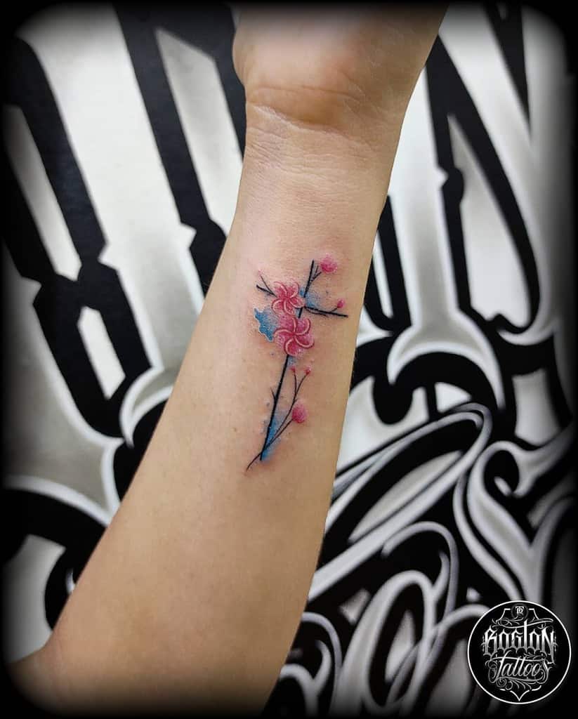 A small and delicate wrist tattoo featuring a cross formed by branches, adorned with soft pink and blue flowers, creating a vibrant and graceful design