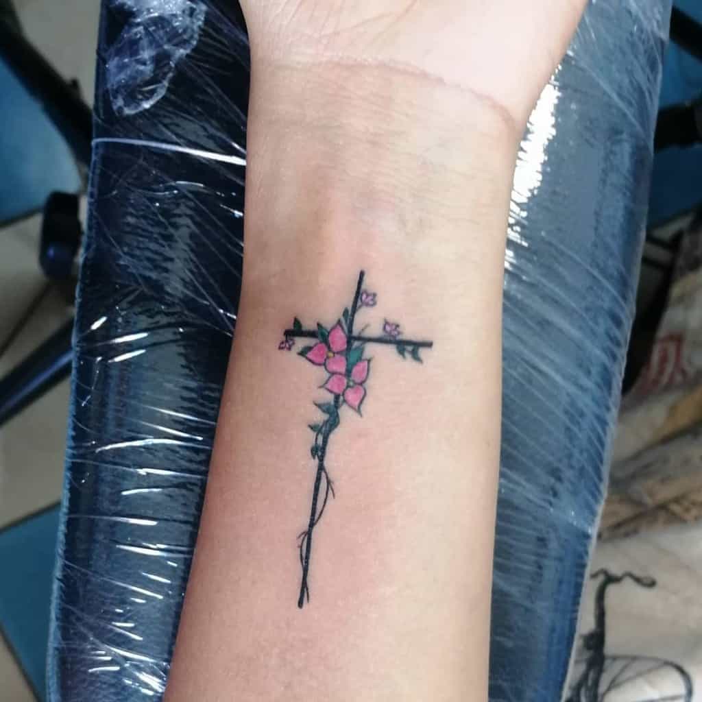 A small wrist tattoo featuring a cross formed by delicate branches, adorned with pink flowers, blending nature and symbolism in a minimalist and elegant design
