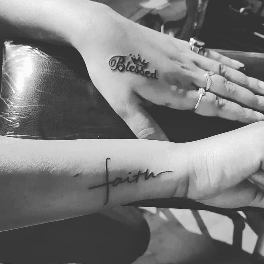 Two hands with tattoos: one reads "Blessed" adorned with a crown, while the other displays "faith" in script, accented by a small cross