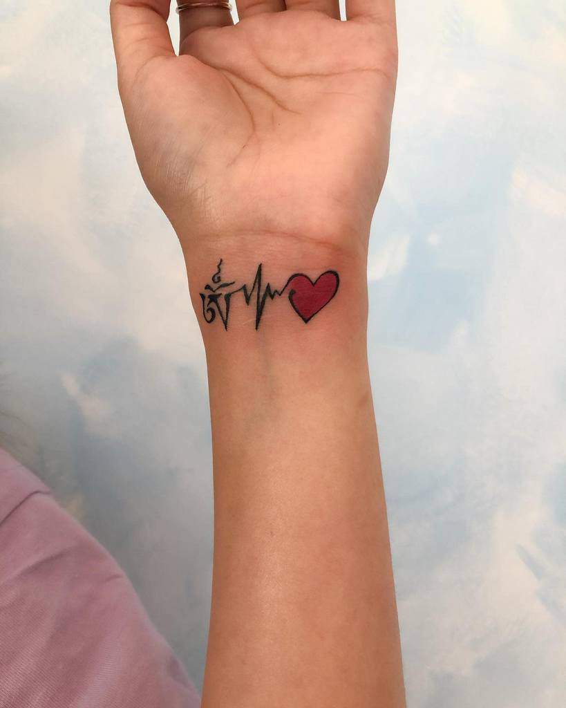 Wrist tattoo featuring a heartbeat line, a red heart, and an intricate symbol next to the heartbeat, representing a mix of faith, hope, and love