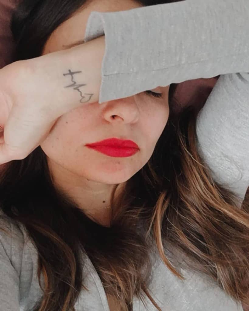 A woman resting with her arm covering her face, showing a minimalistic wrist tattoo of a heartbeat line and heart, symbolizing faith, hope, and love