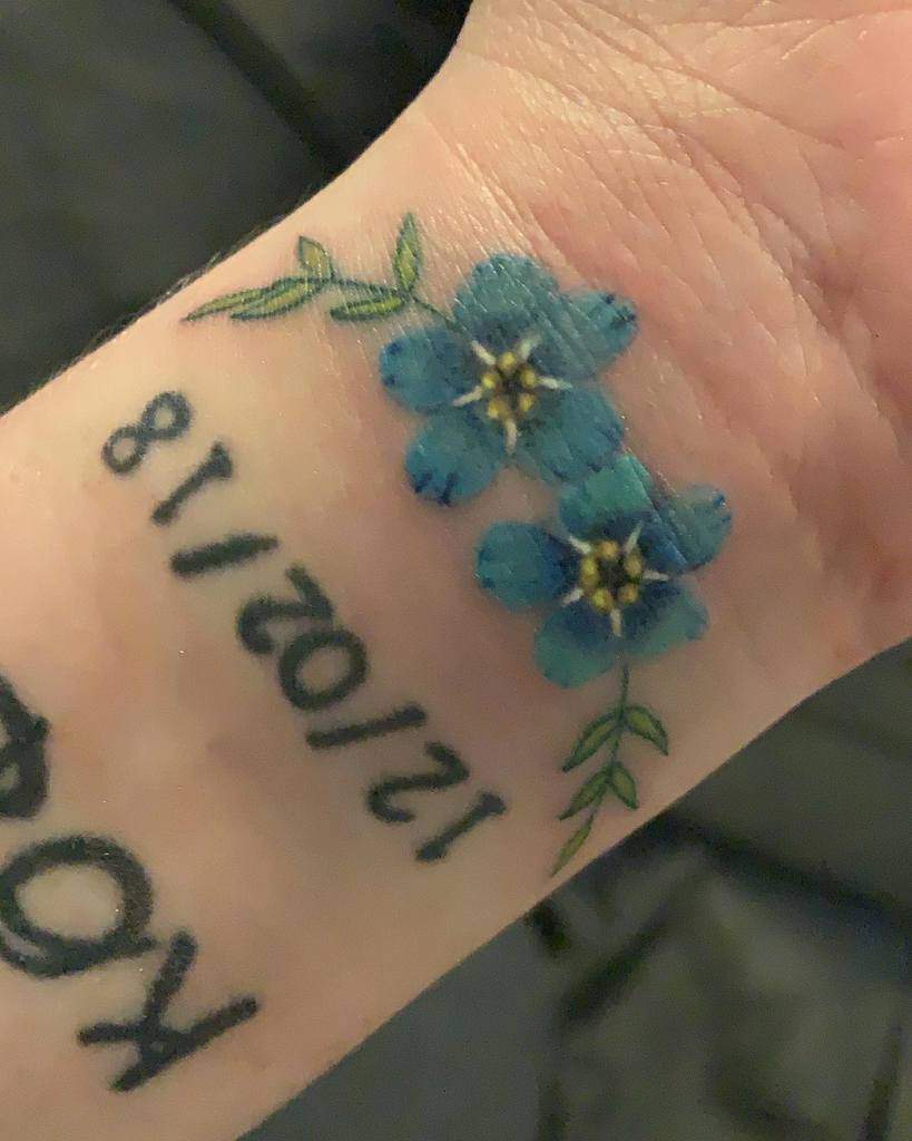 A delicate watercolor tattoo of three blue forget-me-nots with green leaves, placed elegantly on the wrist