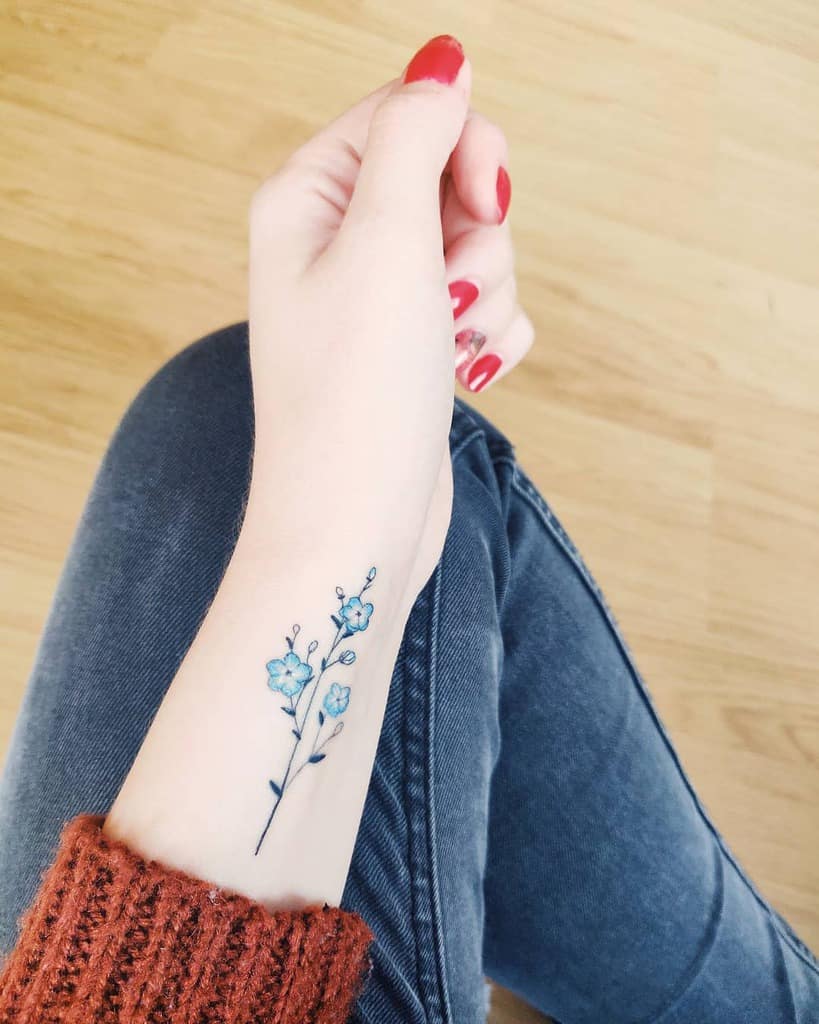 A minimalist tattoo of blue forget-me-nots on a delicate stem, inked on the inner wrist