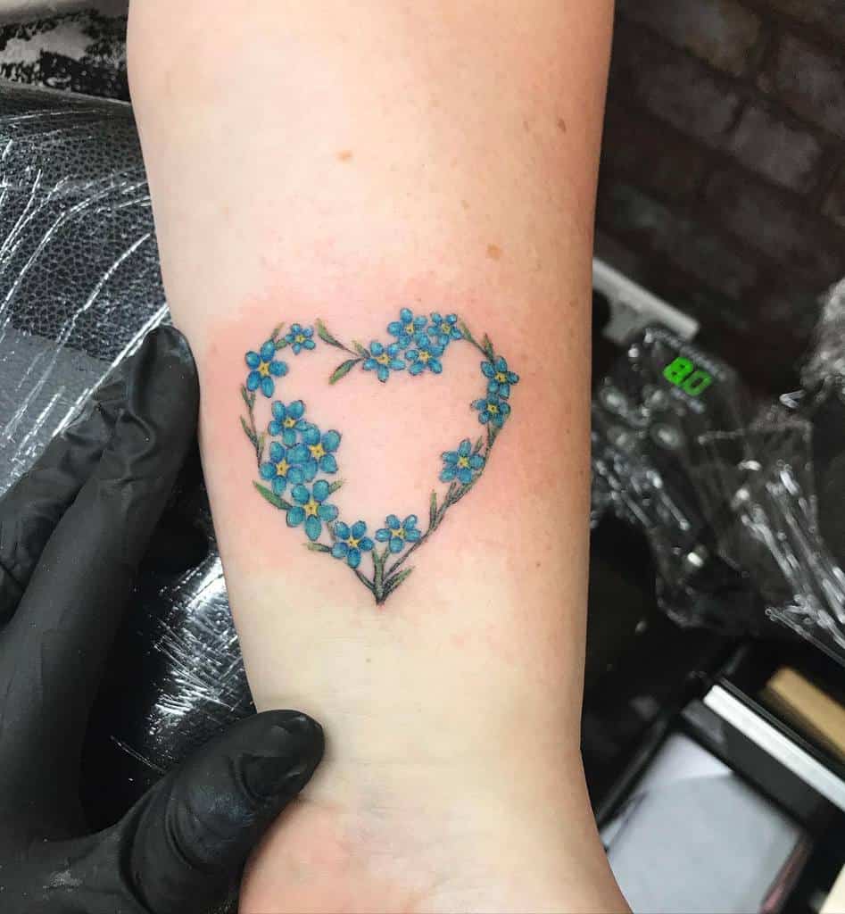 A heart-shaped arrangement of blue forget-me-nots, beautifully tattooed on the inner arm with delicate details and fine linework